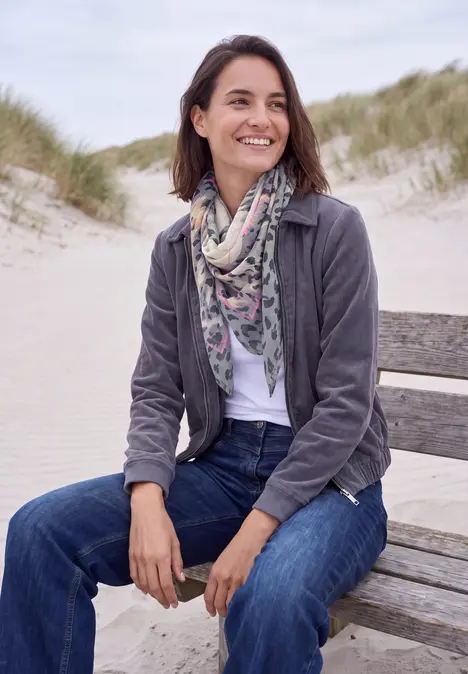 Eine Frau sitzt entspannt auf einer Strandbank, lächelt, trägt Jeans, Schal und Jacke vor Dünen.