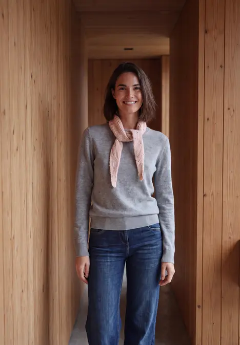 Frau in Holzflur, trägt grauen Pullover, Jeans und helle Seidenschleife, lächelnd zur Kamera.