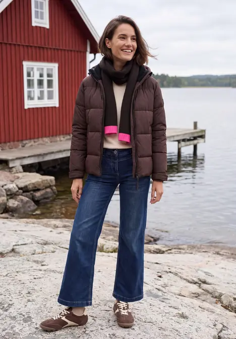 Eine Frau im braunen Steppmantel steht am Wasser neben einem roten Bootshaus.