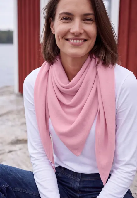 Eine lächelnde Frau in weißem Shirt und rosa Dreieckstuch sitzt am Wasser.