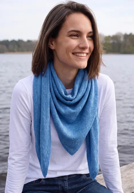Lächelnde Frau mit blauem Dreieckstuch und weißem Shirt sitzt am Wasser, Blick zur Seite.