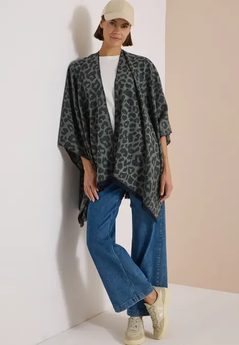 Frau in Cap, grüner Leopard-Poncho, weißes Shirt, Jeans und Sneaker vor hellem Hintergrund