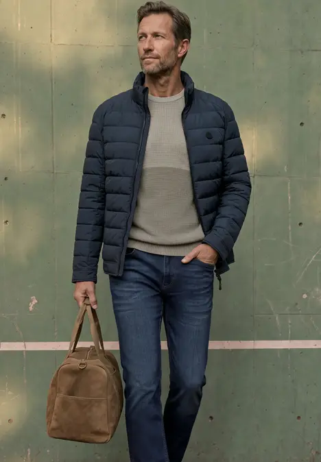 Mann in gesteppter Jacke, Jeans und Pullover trägt eine braune Reisetasche vor grüner Wand.