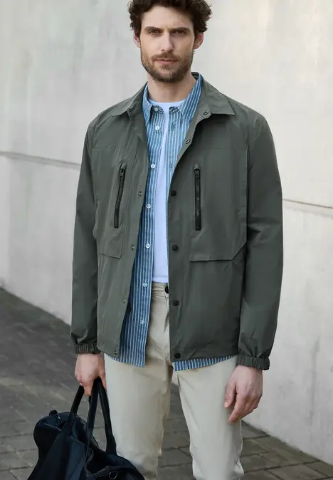 Mann mit grüner Jacke, blauer Hemdjacke und Tasche steht draußen.