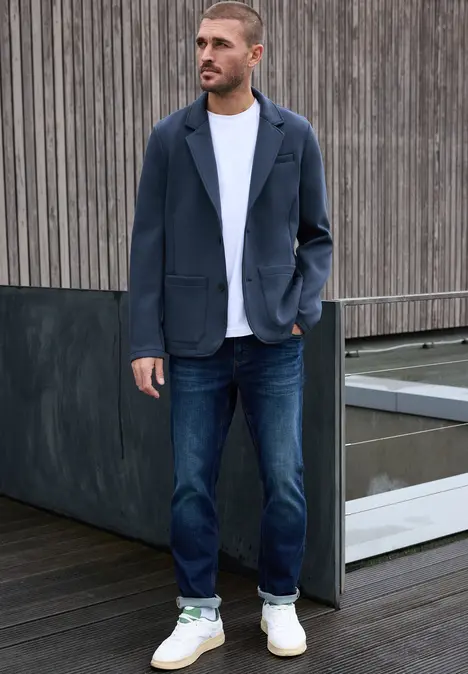 Man in wit T-shirt, donkerblauw colbert, spijkerbroek en witte sneakers voor een houten wand.