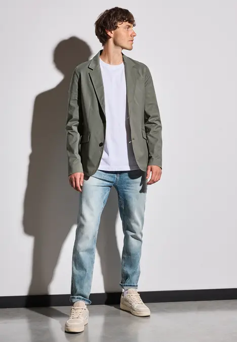 Man in grijs colbert, wit shirt en spijkerbroek voor donkere achtergrond.