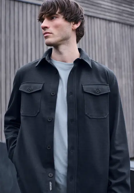 Junger Mann in schwarzer Overshirt und weißem T-Shirt lehnt lässig vor einer Holzverkleidung.