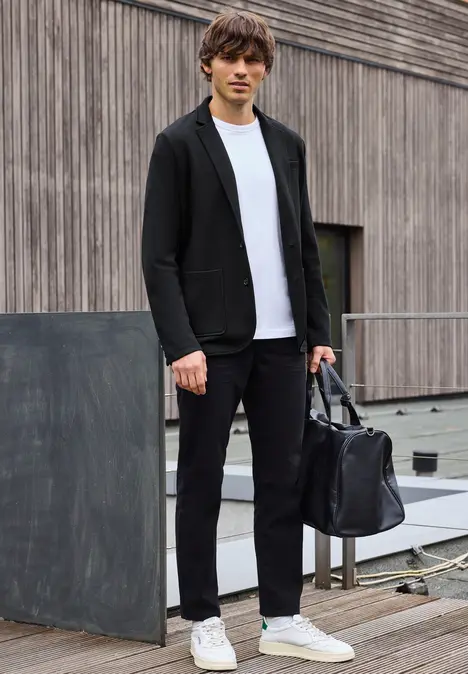 Man in zwarte blazer, wit shirt, sneakers en tas buiten.