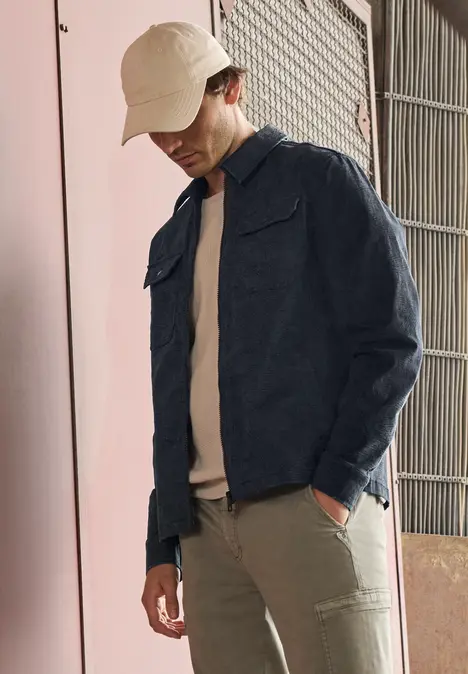 Mann trägt Jeansjacke, beiges T-Shirt, beige Hose und Bucket Hat vor pinker Wand und Metalltor.
