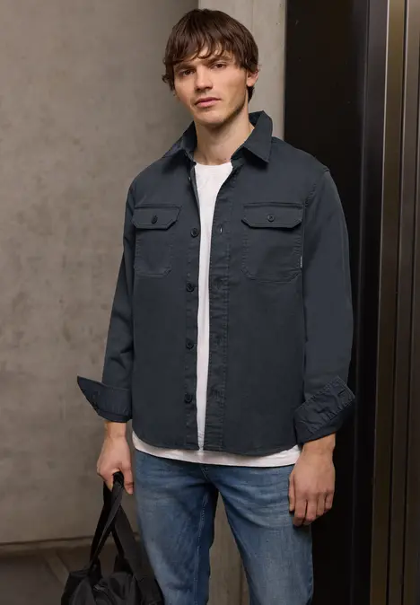 Ein Mann in Jeans, weißem T-Shirt und dunkelgrauem Hemd steht lässig mit einer Tasche in der Hand.