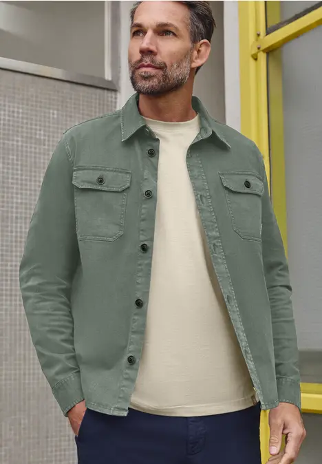 Ein Mann steht lässig in grüner Hemdjacke und weißem T-Shirt vor gelbem Türrahmen.