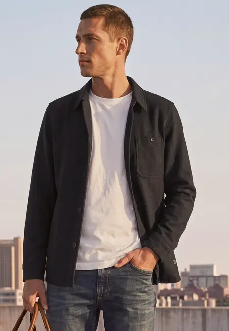 Junger Mann in Jeans, weißem T-Shirt und dunkler Jacke steht vor einer Stadt-Silhouette im Abendlicht.