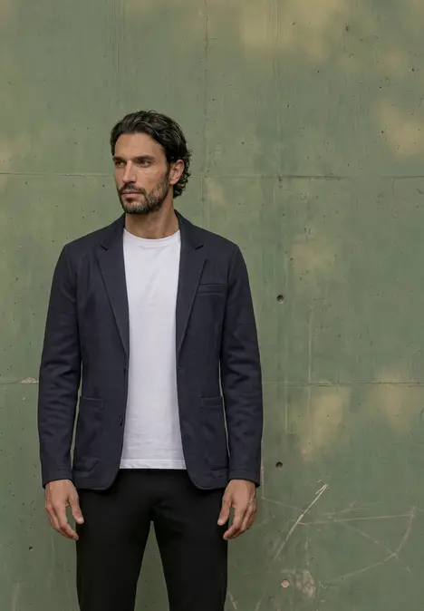 Mann vor grüner Wand trägt dunkelblauen Blazer, weißes T-Shirt und schwarze Hose.