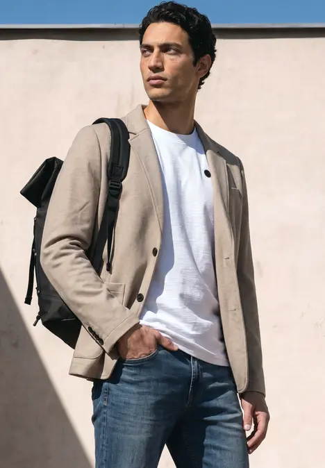 Ein Mann steht vor einer Wand, trägt eine beige Jacke, weißes T-Shirt, blaue Jeans und einen Rucksack.