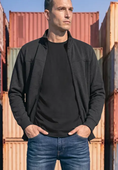 Ein Mann in schwarzem Pullover und Jeans steht vor Stapelcontainern, die Hände in den Taschen, Blick zur Seite.