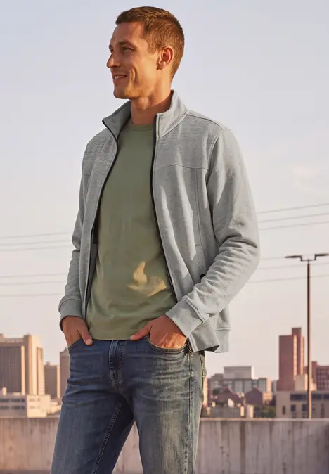 Ein lächelnder Mann in Jeans, grünem T-Shirt und grauer Jacke steht auf einem Dach mit Stadtpanorama im Hintergrund.