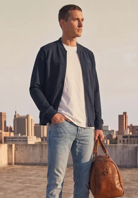 Ein Mann in weißem T-Shirt, schwarzer Bomberjacke und Jeans hält eine braune Ledertasche auf einer Dachterrasse.