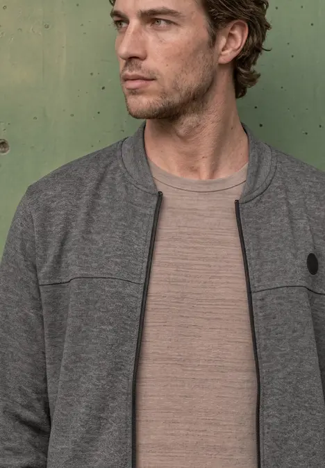 Ein Mann in braunem T-Shirt und grauer Jacke vor grüner Metallwand.