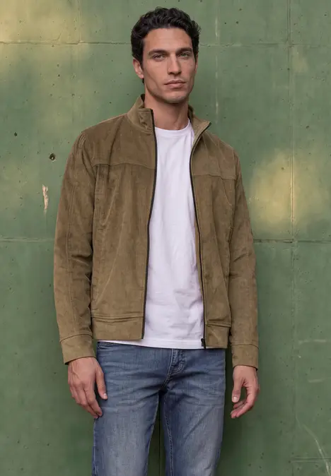 Ein junger Mann in brauner Cord-Jacke, weißem T-Shirt und Jeans steht vor einer grünen Wand.