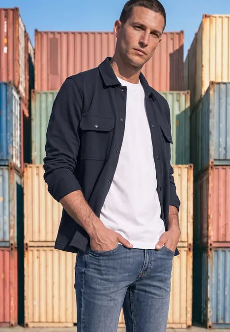Ein Mann in Jeans, weißem T-Shirt und schwarzer Jacke steht lässig vor bunten Containerwänden.