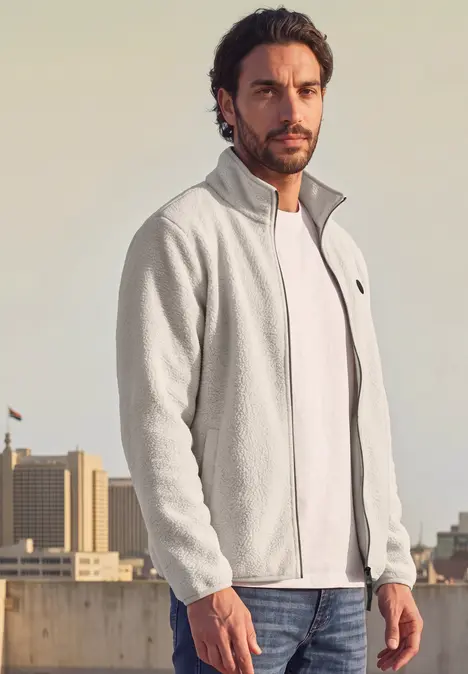 Ein Mann trägt eine graue Fleecejacke über weißem T-Shirt, stehend auf einem städtischen Dach.