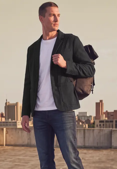 Mann mit Rucksack, grüner Jacke, weißem Shirt und Jeans steht auf einem Dach vor einer Stadtkulisse.