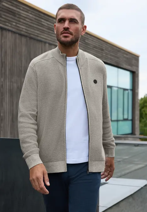 Ein Mann steht vor einem modernen Gebäude und trägt eine bequeme, hellgraue Zip-Jacke über weißem T-Shirt.