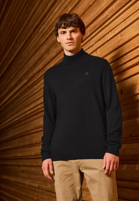 Mann in schwarzem Pullover und beiger Hose vor Holz-Wand.