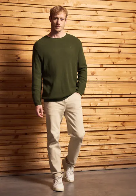 Mann in grünem Pullover und beiger Hose steht vor Holzwand.