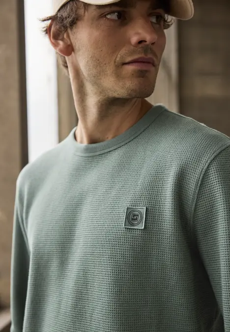Mann trägt grünen, strukturierten Pullover und steht in einem hellen Raum vor einer Backsteinwand.