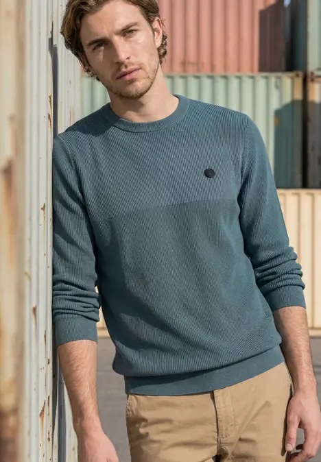 Ein Mann in türkisfarbenem Pullover und beiger Hose steht vor bunten Containern im Freien.