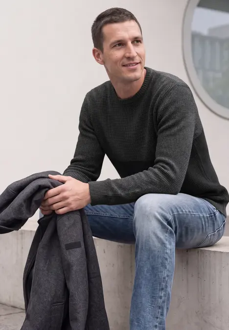 Ein Mann sitzt vor einem Gebäude, trägt Jeans und Pullover, hält eine Jacke und blickt seitlich.