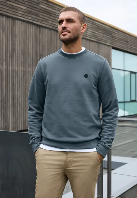 Ein junger Mann in blauem Sweatshirt und beiger Hose steht vor modernem Gebäude mit Holzwand und Glasfront.