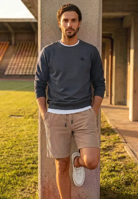 Mann lehnt lässig an Säule im Freien, trägt Sweatshirt, Shorts und weiße Sneaker.
