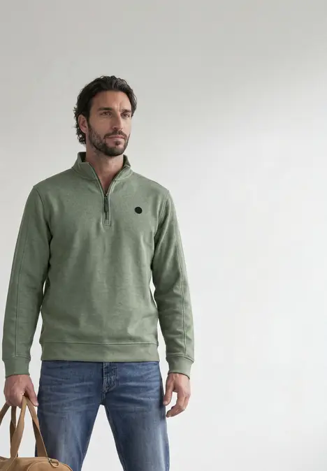 Ein Mann mit Bart trägt einen olivgrünen Pullover und Jeans, steht vor hellem Hintergrund und hält zwei Beuteltaschen.
