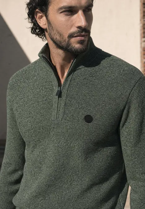 Mann mit dunkelgrünem Strickpullover mit halbem Reißverschluss und Logo, schaut seitlich.