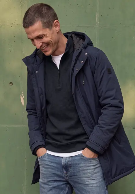 Homme en jean, pull et parka, souriant, appuyé contre un mur vert.