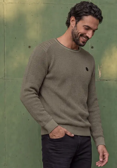 Ein lächelnder Mann trägt einen olivgrünen Pullover und schwarze Hose vor grünem Hintergrund.