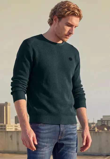 Ein junger Mann steht auf einer Dachterrasse und trägt einen schwarzen Pullover und blaue Jeans.