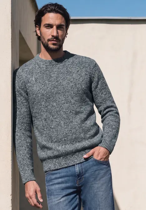 Ein Mann lehnt lässig an einer Wand, trägt grauen Pullover und Jeans