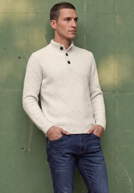 Homme devant un mur vert, en pull clair et jean.
