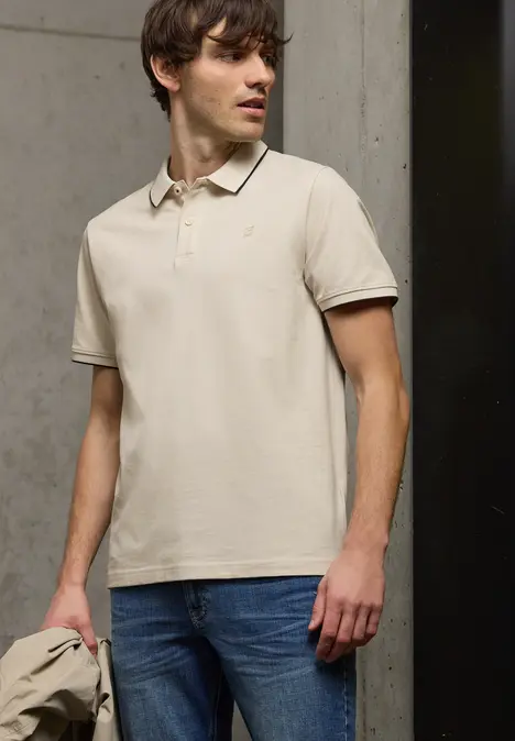 Ein Mann trägt ein beiges Poloshirt und blaue Jeans und hält eine passende Jacke vor neutralem Hintergrund.