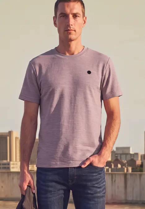 Een man in spijkerbroek en T-shirt staat voor een stedelijke achtergrond en houdt nonchalant een tas vast.