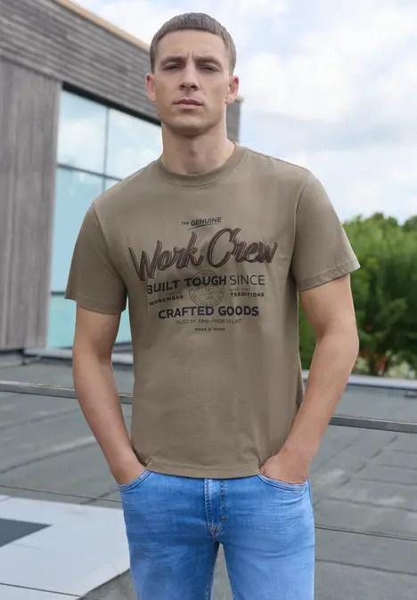 Een jonge man op een dakterras, met een kaki t-shirt met print en een blauwe spijkerbroek.