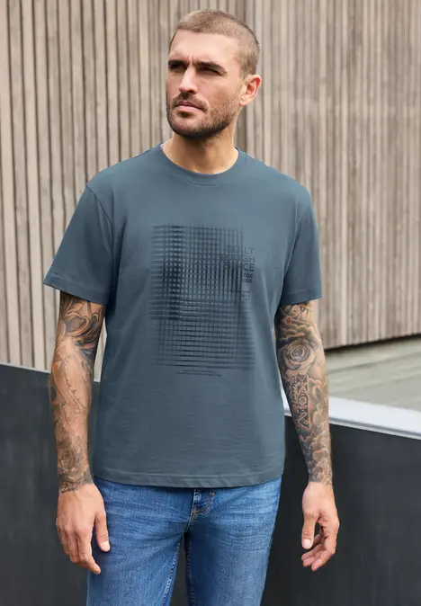 Un homme tatoué en t-shirt et jean se tient devant un mur en bois et regarde sur le côté.