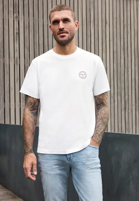 Een getatoeëerde man draagt een wit t-shirt en spijkerbroek, staand voor een houten wand.
