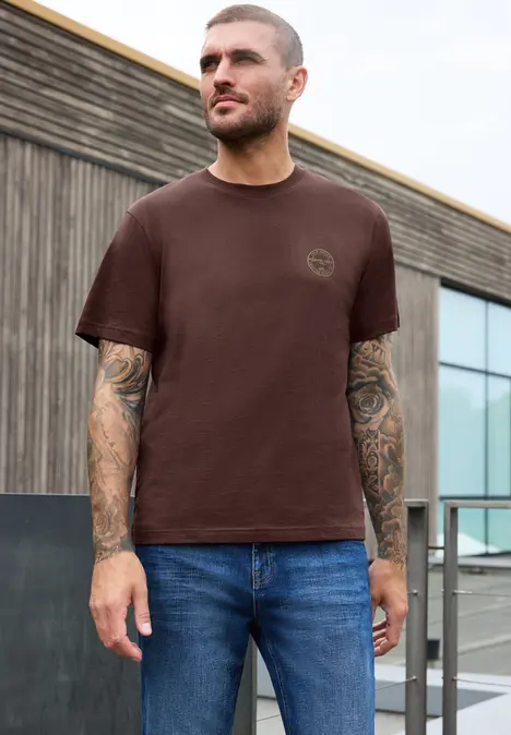 Junger Mann mit braunem T-Shirt und Jeans, tätowierte Arme, steht vor modernem Gebäude.