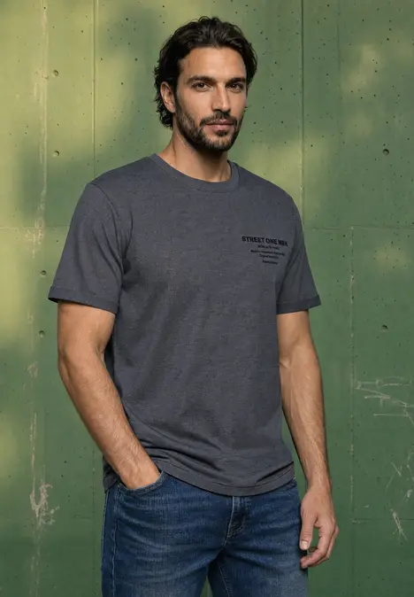 Ein Mann mit dunklem Haar und Bart trägt ein graues T-Shirt und Jeans vor einem grünen Hintergrund.