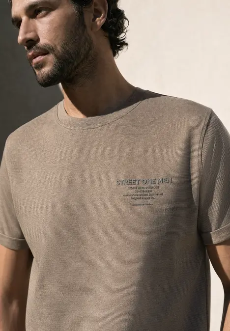 Un homme de profil porte un t-shirt beige avec un discret lettrage sur la poitrine.