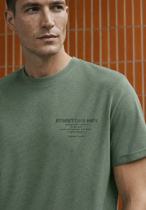 Man in groen T-shirt met klein logo, staat voor oranje betegelde muur.